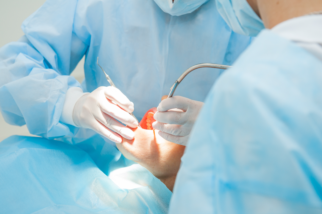 Dentists in blue protective gowns and gloves performing dental examination with dental tools inside a patient's mouth.